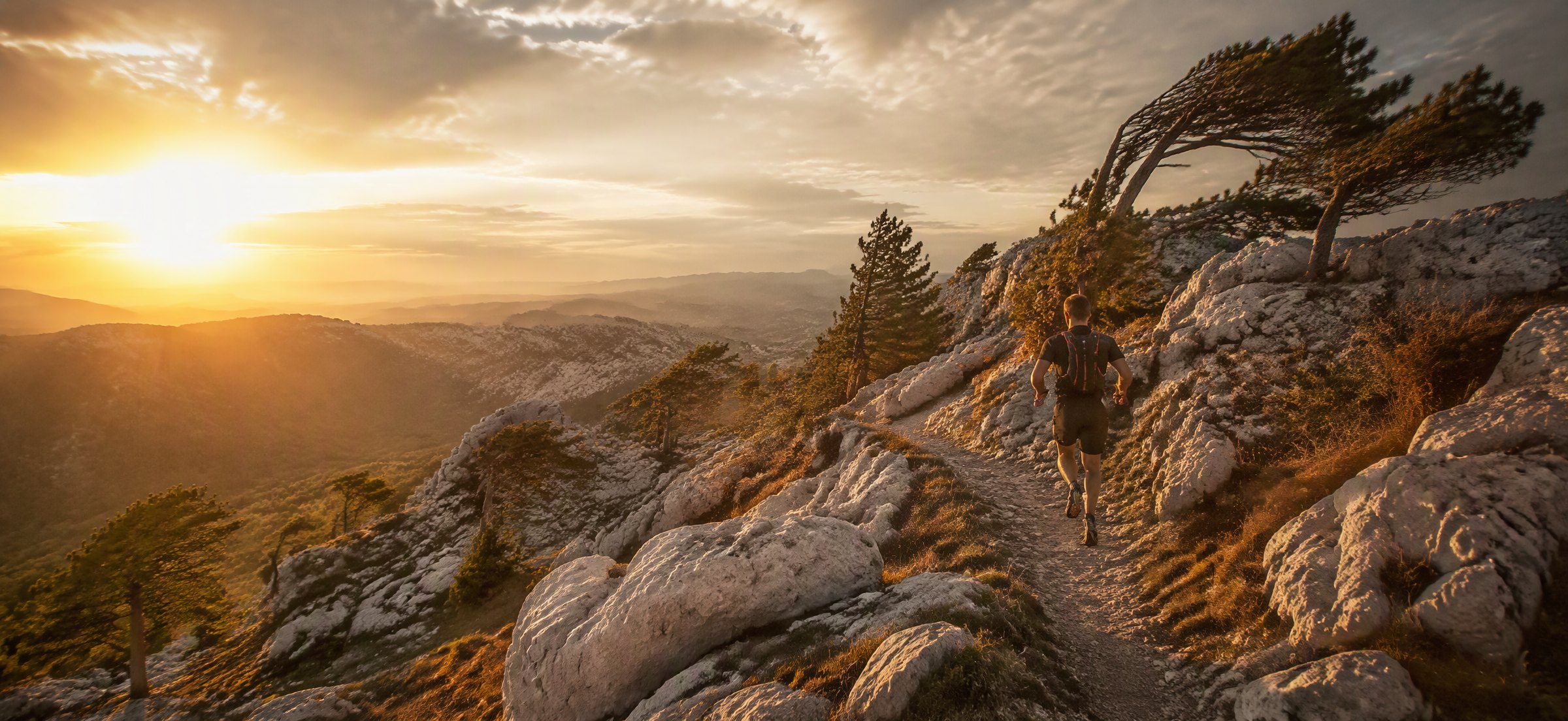 Trail runner on a rocky karst ridge at sunrise, wind-bent pines and the vast plateau below